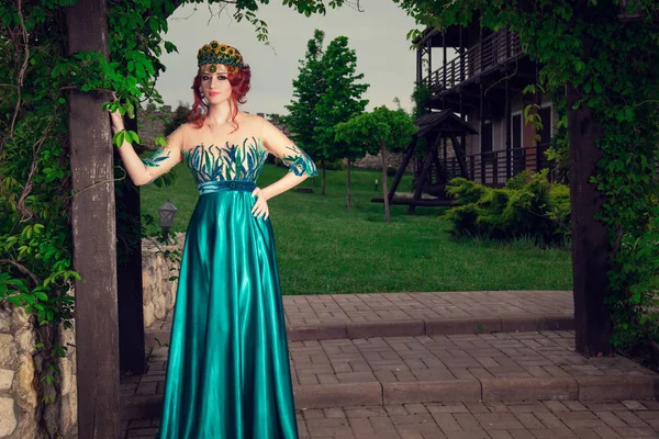 Woman with crown on head in green long dress posing looking at you camera in a beautiful garden in the evening. Bridal gown, makeup and hair style concept.
