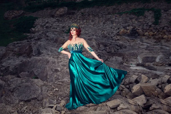 Woman in full body with crown on head in green long dress posing looking at you camera in a beautiful stone cascade in the evening. Bridal gown, makeup and hair style concept.
