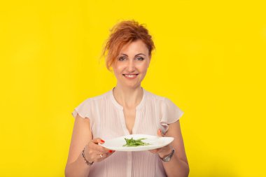Sarı arka planda izole yeşil salata bir tabak ile mutlu gülümseyen olgun kadın. Diyet vejetaryen veya ham vegan kavramı.