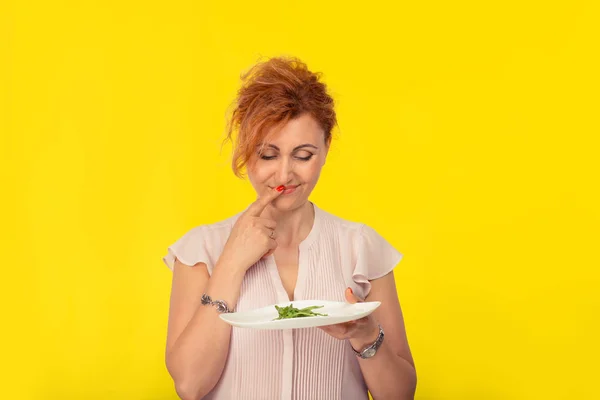 Unhappy mature woman with a plate of green salad looking at it with disgust unhappy to eat it, thinking about meat isolated on yellow background. Dieting vegetarian or raw vegan concept.