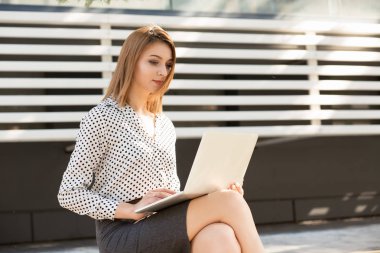 Gülümseyen genç kadın bir şehir sokak arka planda dışında onun laptop kullanarak. Uzun bob saç kesimi ile resmi giyim ofisi veya kolej yakınında açık havada oturan kız