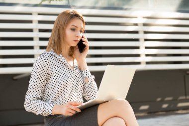 Sevimli genç kadın bilgisayar da çalışırken cep telefonu konuşuyor, bir yaz gününde dizüstü bilgisayar açık havada. Uzun bob saç kesimi ile resmi giyim ofisi veya kolej yakınında açık havada oturan kız