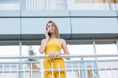 Büyüleyici genç samimi kadın, rahat sarı tulum öğrenci cep telefonu onun ofis, kolej, arka planda üniversite binasının metalik balkonda açık havada duran konuşurken