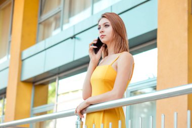 Büyüleyici genç samimi kadın, rahat sarı tulum öğrenci cep telefonu onun ofis, kolej, arka planda üniversite binasının metalik balkonda açık havada duran konuşurken