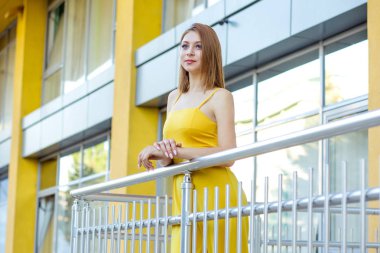 Güzel kadın ayakta, bir kentsel modern binanın balkonunda poz yan daydreaming bakıyor. Uzun saçlı sarı tulumlu Düşünceli Kız, bob saç kesimi.