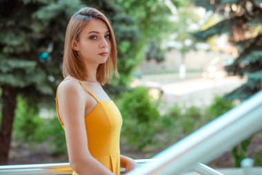 Güzel kadın ayakta, bir kentsel modern metalik binanın balkonunda size bakarak poz, uzun saçlı sarı tulum elbiseli kamera Düşünceli Kız, bob saç kesimi.