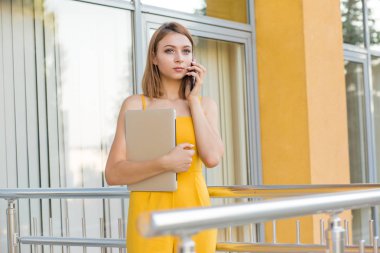Büyüleyici genç kadın, gündelik sarı tulum öğrenci cep telefonu konuşurken ve diğer taraftan açık havada ofis ayakta bilgisayar tutarak, kolej, arka planda üniversite binası