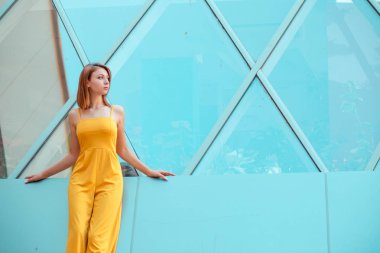 Güzel kadın ayakta, yan daydreaming bakarak bir arka plan olarak aynalı cam pencerelerden bir kentsel modern binada poz. Uzun saçlı sarı tulumlu Düşünceli Kız, bob saç kesimi.
