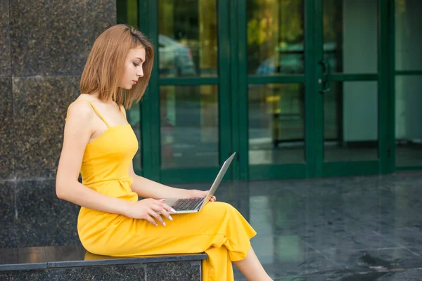 Side profile portrait of woman sitting outside, using laptop. Girl standing outdoors near a green building made of glass in a yellow jumpsuit with long hair, bob haircut.