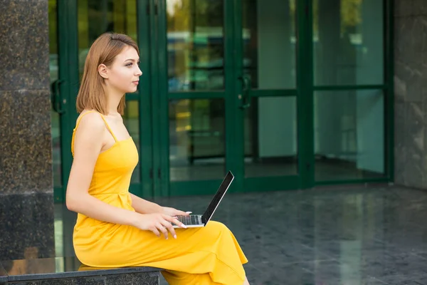 Side profile portrait of woman sitting outside holding opened laptop looking to side thoughtful. Girl standing outdoors near a green building made of glass in a yellow jumpsuit with long bob haircut