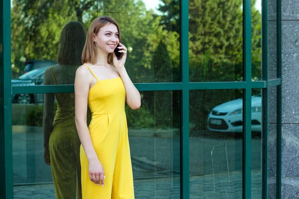 携帯電話で話す女性は幸せ 長い髪の黄色のジャンプスーツ ボブヘアカットでガラスで作られた緑の建物の近くに屋外に立っている女の子 — ストック写真