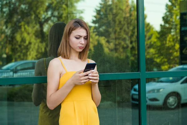 Upsetting message. Front view of a serious sad depressed girl checking a phone message standing near the office glass door. Mixed race modern model Girl in a yellow jumpsuit with long hair, bob haircut.