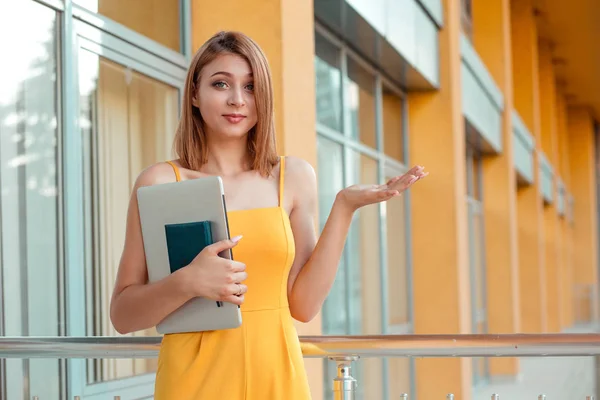 笑顔の女性 無関心を示す学生 黄色のジャンプスーツを着て オフィスや大学の建物の近くに立ってノートパソコンや本を手に持つ手で何のジェスチャー — ストック写真