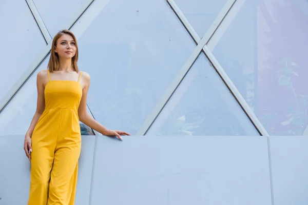 Beautiful woman standing, posing on an urban modern building from mirrored glass windows as a background looking to the side daydreaming. Thoughtful Girl in a yellow jumpsuit with long hair, bob haircut.