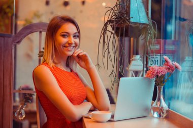Dizüstü bilgisayarının önünde oturan ve kameraya bakıp gülümseyen başarılı bir kadın. Mutfağında, pencerenin yanında ya da kahve dükkanında gülüyor. Beyaz benekli kırmızı elbise giyen modaya uygun bir kafe..