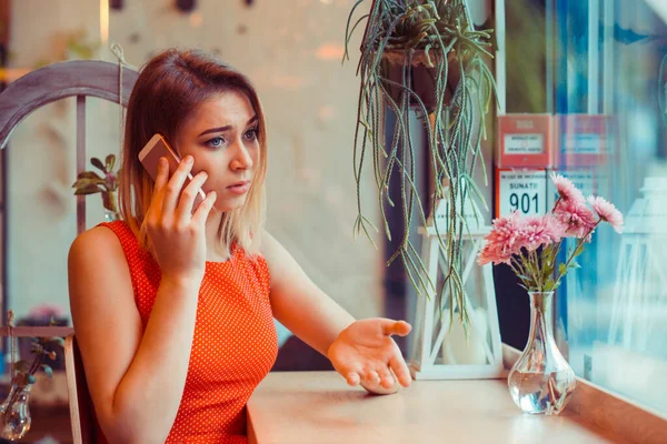 窓の近くの自宅のキッチンやコーヒーショップでテーブルに座っている間 電話で話す不幸な心配の不満女性は 流行のカフェ赤いドレスを着て 否定的な感情の表情感 — ストック写真