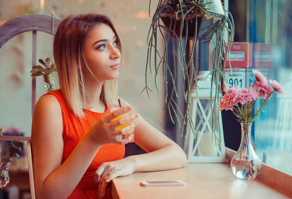 カフェシティライフスタイル思慮深い女性がオレンジジュースを飲んで ガラス窓から通りを外を見て 屋内ジュースバーに座って 家庭用キッチンやコーヒーショップで 流行のカフェ赤いドレスを着て — ストック写真