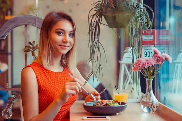 Healthy eating, dieting and people concept. Closeup portrait young woman eating vegetable salad looking at you camera smiling in home kitchen, near window or in a coffee shop, trendy cafe in red dress