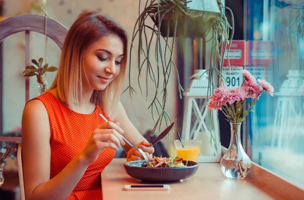 Sağlıklı beslenme, diyet ve insanların konsepti. Yakın plan, sebze salatası yiyen genç bir kadın ev mutfağında, pencerenin yanında ya da kahve dükkanında gülümseyen kameraya bakıyor. Kırmızı elbiseli şık bir kafe.