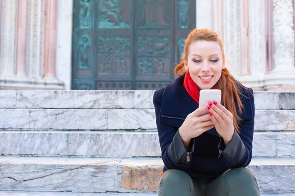 Woman received a good text message on her cell phone while sitting on stairs in the city. Girl texting on smartphone wearing blue coat, red scarf, redhead long hair