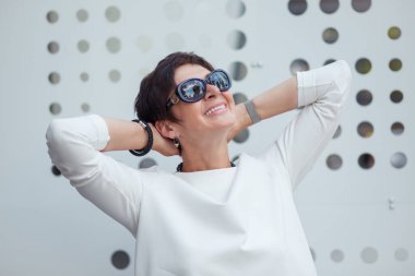 50 yaşında olgun bir kadın olarak gülümseyen, yaz tatili hayali kuran, güneş gözlüklü, kafasının arkasında duran, beyaz metalik duvarın yanında duran bir kadın. Mutlu bayan rahatlatıcı Hoş anılar konsepti