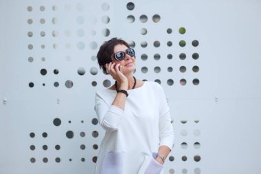 Beyaz bluz ya da elbise ve siyah güneş gözlüğü giyen modaya uygun orta yaşlı bir kadın akıllı telefonuyla konuşuyor. Metalik bir standın yanında neşeli bir şekilde duruyor..