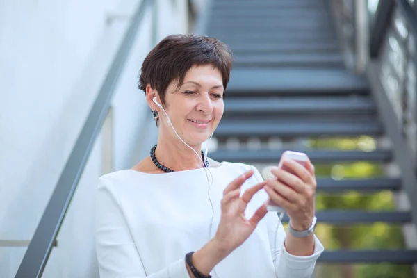 Happy woman holding mobile phone in ear-pods standing near a metallic fence stairs looking at phone serious. Short hair brunette 50 years old, lady in white blouse shirt using cell listening to music.