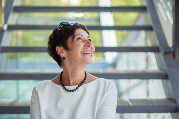 Cheerful stylish middle-aged woman sitting on stairs on street relaxing daydreaming looking up to the sky or copyspace and smiling happily wearing white blouse dress and black pearls fill on neck.