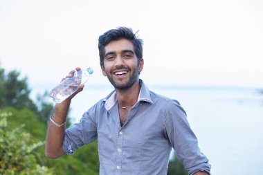 Biraz su iç. Profilde gülümseyen bir adam su şişesinden içmek üzere olan izole edilmiş doğa ağaçları ve deniz altyapısı.