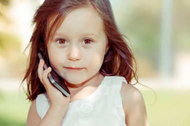 Akıllı telefonu olan bir çocuk. Cep telefonuyla annesiyle konuşan komik bir kızın yakın plan portresi. Babası yaz arka planında yeşil ağaçları izole etmiş. Çocuklar ve teknoloji konsepti