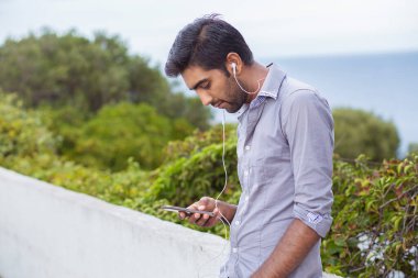 Bir kısa mesaj alınıyor. Gri tişörtlü yakışıklı adam dışarıda Mobil Akıllı Telefona bakıyor, yeşil çalılık, arka planda deniz.