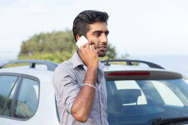 Cep telefonuyla konuşan, arabasının kapısına dayanmış, sohbet eden bir adam.