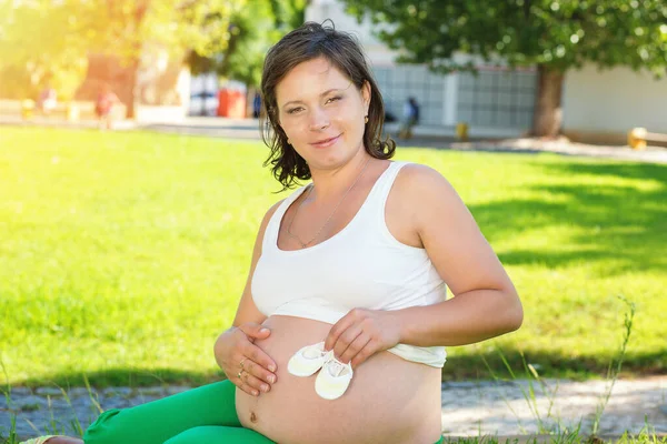 Pregnant Woman touching her belly, tummy playing with little baby shoes sitting outdoors on a green grass meadow in a city park on a sunny day. Happy mom to be, Healthy Pregnancy concept Parenthood