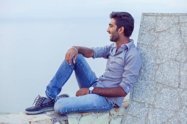 Mavi gökyüzüne gülümseyen bir adam denizin üzerindeki beton bir duvar köprüsünde oturmuş özgürlüğünü kutlarken derin bir nefes alıp mutlu mesut nefes alıyor. Olumlu insan, barış zihni konsepti. Özgür mutlu adam günbatımının tadını çıkarıyor