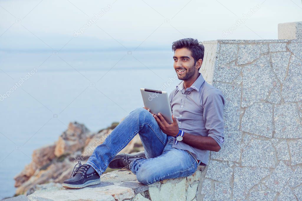 Man smiling happy while using pad looking to blue sky while sitting on a concrete bridge above the sea taking deep breath enjoying freedom at sunset sea on background. Cheerful excited cool person