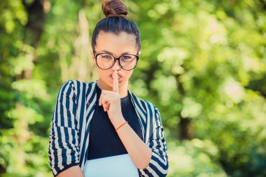 Dudaklarında işaret parmağı olan genç, güzel esmer bir kadın. Sessizlik işareti gösteriyor. Arka planda yeşil bir parkta..