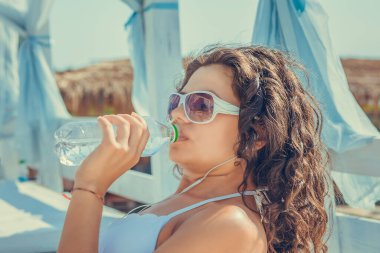 Yazın dışarıdayken su iç. Güzel görünümlü, güneş gözlüklü, Afro saçlı, beyaz mayo giymiş, güneşin tadını çıkaran, gölde izole edilmiş bir şişe su içen bir kadın.