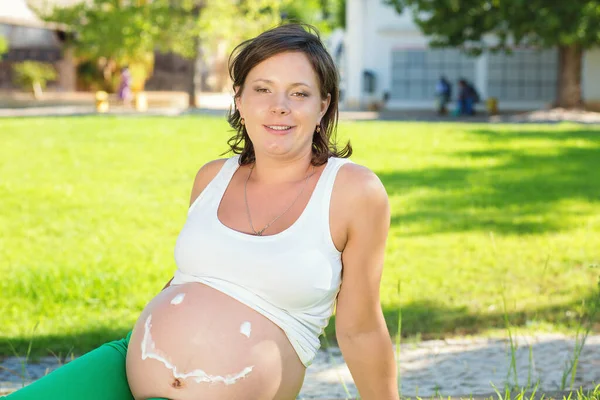 Belly of pregnant happy woman  with cream in shape of face with smile, woman applying moisturizing cream for stretch marks while sitting in park on green grass meadow outdoors. Pregnancy and skin care concept
