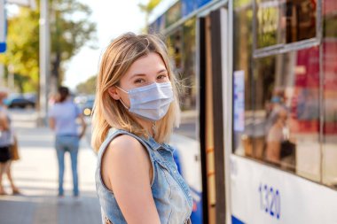 Cheerful woman wearing a sterile protective medical mask against coronavirus, Covid-2019 Asian pandemic sars virus at public bus station in European city street looking at you camera people background clipart
