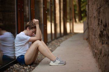 Düşünceli ve üzgün bir kız. Kısa saçlı bir kadının portresi. Yazın arka planda cam bir duvarın yanında yerde otururken üzüntüyle düşünüyor. Yatay resim.