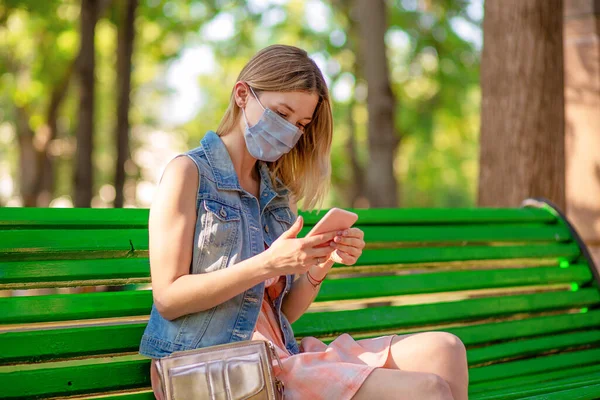 Woman in a park sitting on a bench alone, looks at her phone while in her protective mask using app looking to avoid sick people. Social distancing and quaratine durig Covid-19 pandemic
