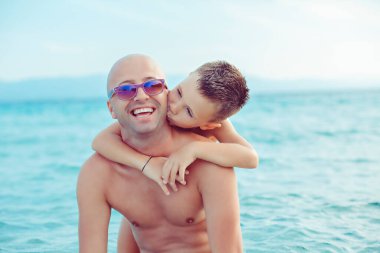Mutlu baba, mavi okyanus suyunun arka planında onu kucaklayan oğlunun öpücüğünden zevk alır..