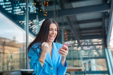 Haberler iyi. Heyecanlı mutlu Hintli kız çevrimiçi zaferi kutluyor, telefonu tutuyor, kameraya bakıyor, balkonda oturuyor, çok mutlu, başarılı genç bir kadın zaferi kutluyor, zafer kazanıyor, gülüyor.