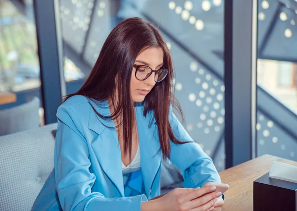 Woman looking wireless to a music video on a smartphone with headphones at her home balcony or in a coffee shop, trendy cafe wearing formal blue suit. Multicultural model.