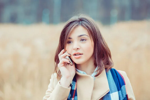 Portrait of a beautiful frustrated woman talking on the mobile phone in a park with a light brown unfocused park nature background