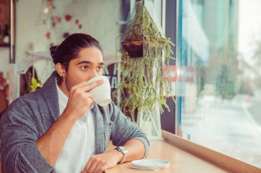 Man enjoying drinking his cup of tea coffee, hot beverage. Closeup portrait of a handsome guy wearing formal white shirt and gray blouse sitting near window at a table in living room or coffee shop.
