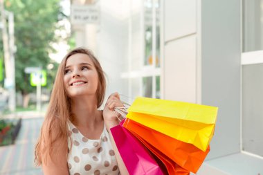 Alışveriş yaptıktan sonra sokakta yürürken gülümseyen ve bakışlarını çeviren parlak kağıt torbalı çekici genç bir kadın.