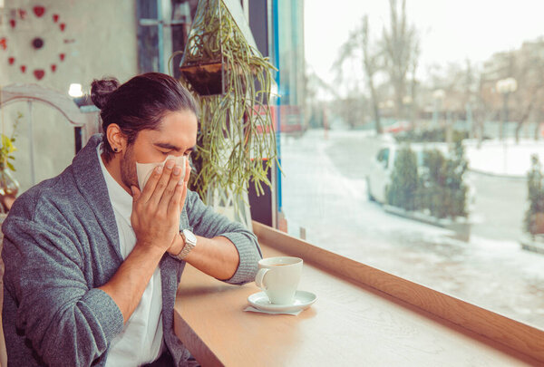 Flu. Young man got nose allergy, flu sneezing nose sitting at the table in a trendy cafe coffee shop with a cup of hot beverage beside. Closeup portrait of a multicultural guy wearing casual clothing