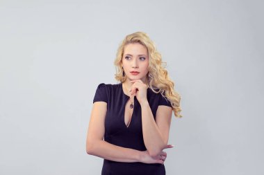 Şık sarışın kadın, derin derin düşünürken çenesine dokunuyor. Beyaz iş adamı, siyah resmi elbise giymiş, uzun sarı kıvırcık saçlı, açık gri arka planda, kopyalama alanı olan açık gri bir stüdyo.