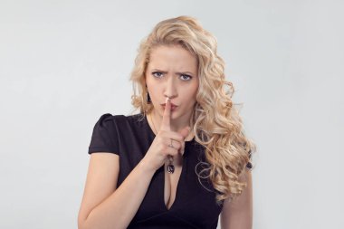 Hush, shh. Modern serious angry young blond woman in dress and glasses holding finger on lips asking to keep silence or a secret. Caucasian Business person in black formal dress, long blond curly hair